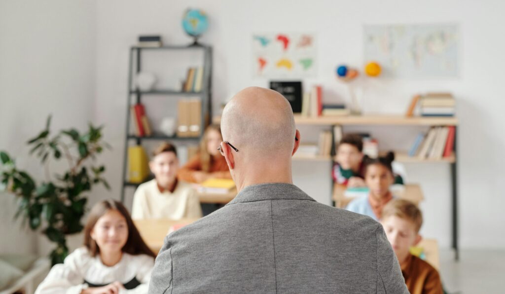 pexels-photo-5211428-5211428 Bald teacher in a classroom engaging with attentive students sitting at desks.